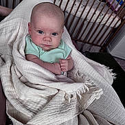 Lillie is registered to the contest to win money with this photo: baby, blanket, infant, wrapped, crib, wooden_crib, blue_eyes, clothing, person, indoor, soft_texture, fringe, bedroom, toy, serious_expression, human, newborn, portrait, child, resting