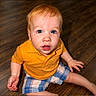 toddler, child, blue_eyes, orange_shirt, plaid_shorts, sitting, wooden_floor, indoor, cute, curious, baby, person, portrait, casual_clothing, young_child, looking_up, small_hands, barefoot, bright_colors, playful