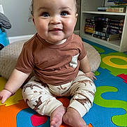 Calais is registered to the contest to win money with this photo: baby, blue_eyes, books, bookshelf, brown_shirt, child, colorful_mat, curly_hair, cushion, cute, dinosaur_pants, feet, floor, hand, indoors, person, playroom, sitting, smiling, young