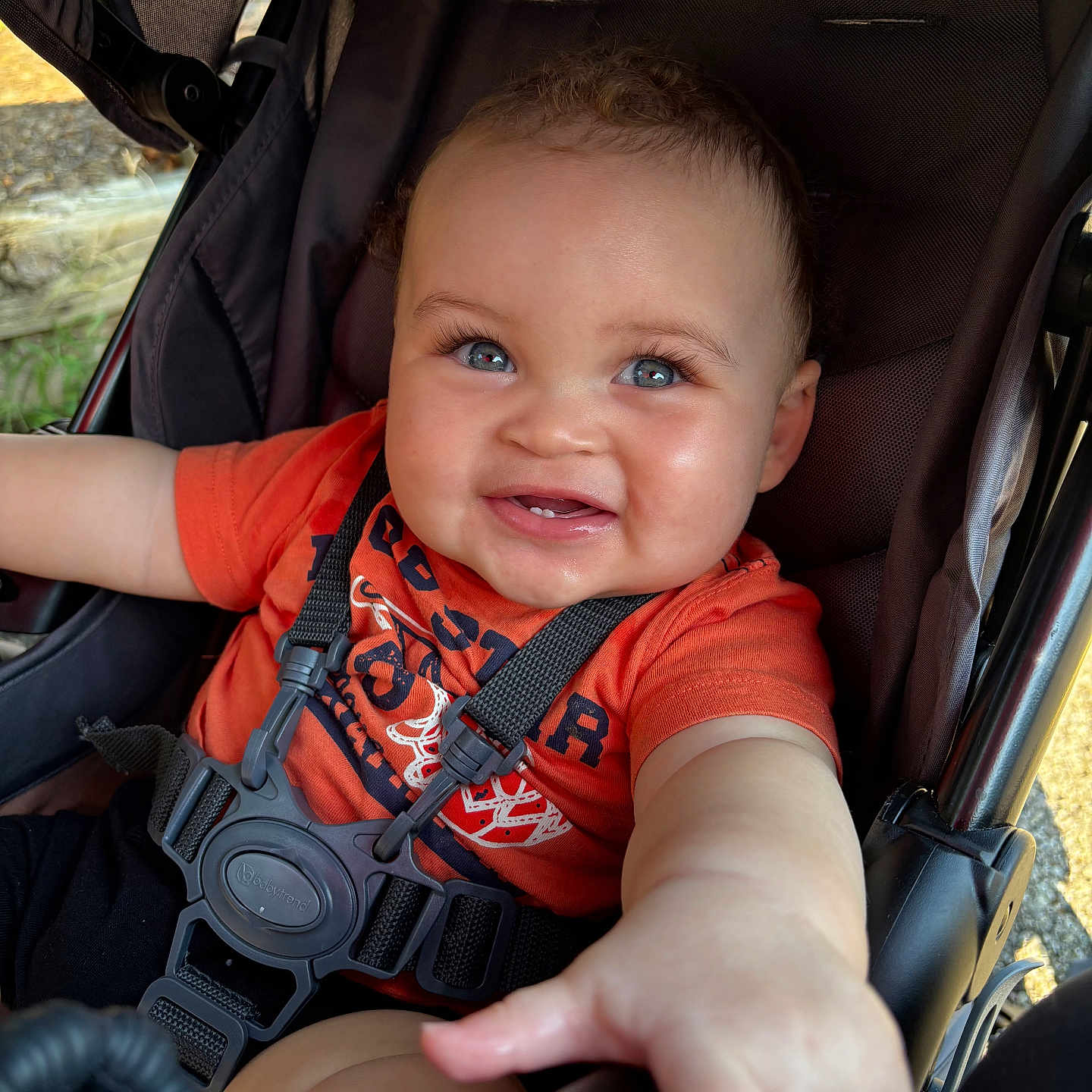 Calais is registered to the contest to win money with this photo: baby, black_harness, blue_eyes, cheeks, child, closeup, curly_hair, cute, happy, infant, natural_light, orange_shirt, outdoor, person, portrait, reaching_hand, safety_strap, seat, smiling, stroller
