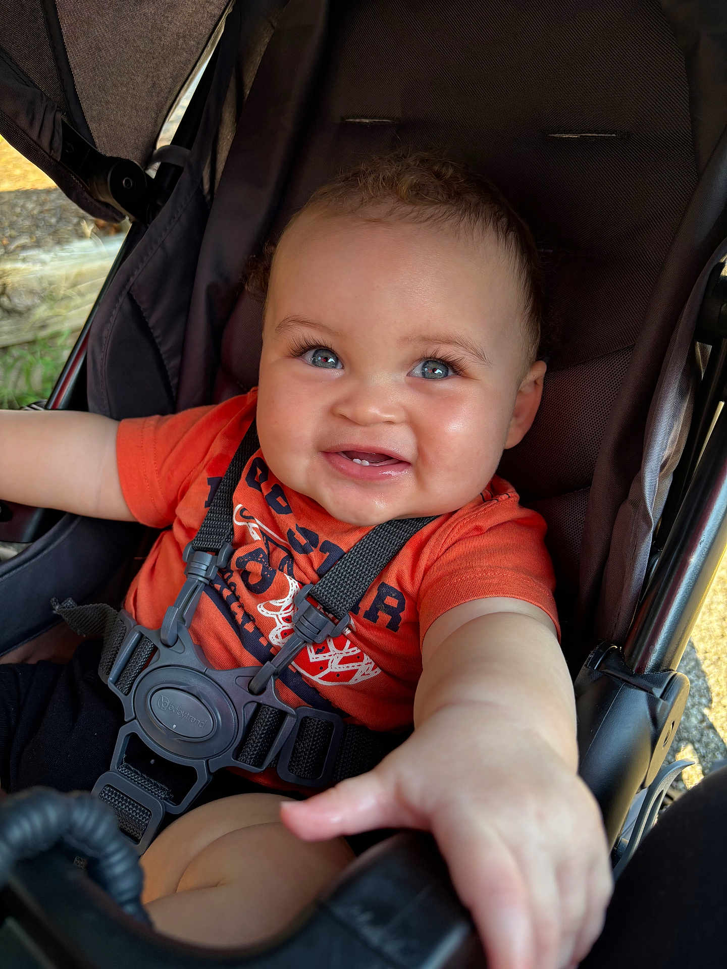 Calais is registered to the contest to win money with this photo: baby, child, stroller, orange_shirt, black_harness, blue_eyes, smiling, reaching_hand, curly_hair, outdoor, natural_light, cheeks, infant, seat, safety_strap, portrait, cute, happy, person, closeup