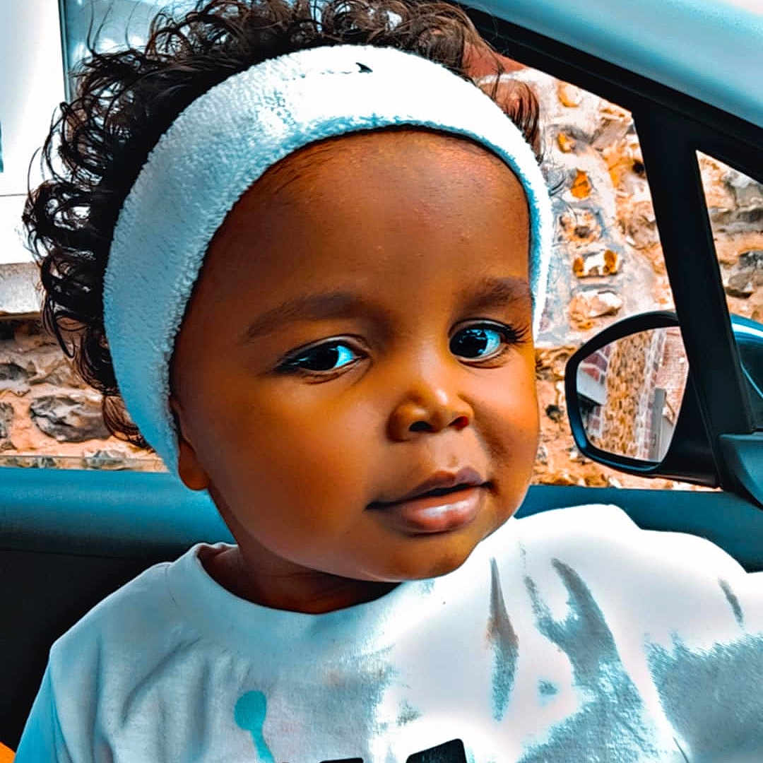 Kaïss participe au concours pour gagner de l'argent avec cette photo : black_hair, car_interior, casual_clothing, child, closeup, curly_hair, cute, expression, face, graphic_print, headband, indoors, person, portrait, sitting, stone_wall, toddler, white_tshirt, window, young_child