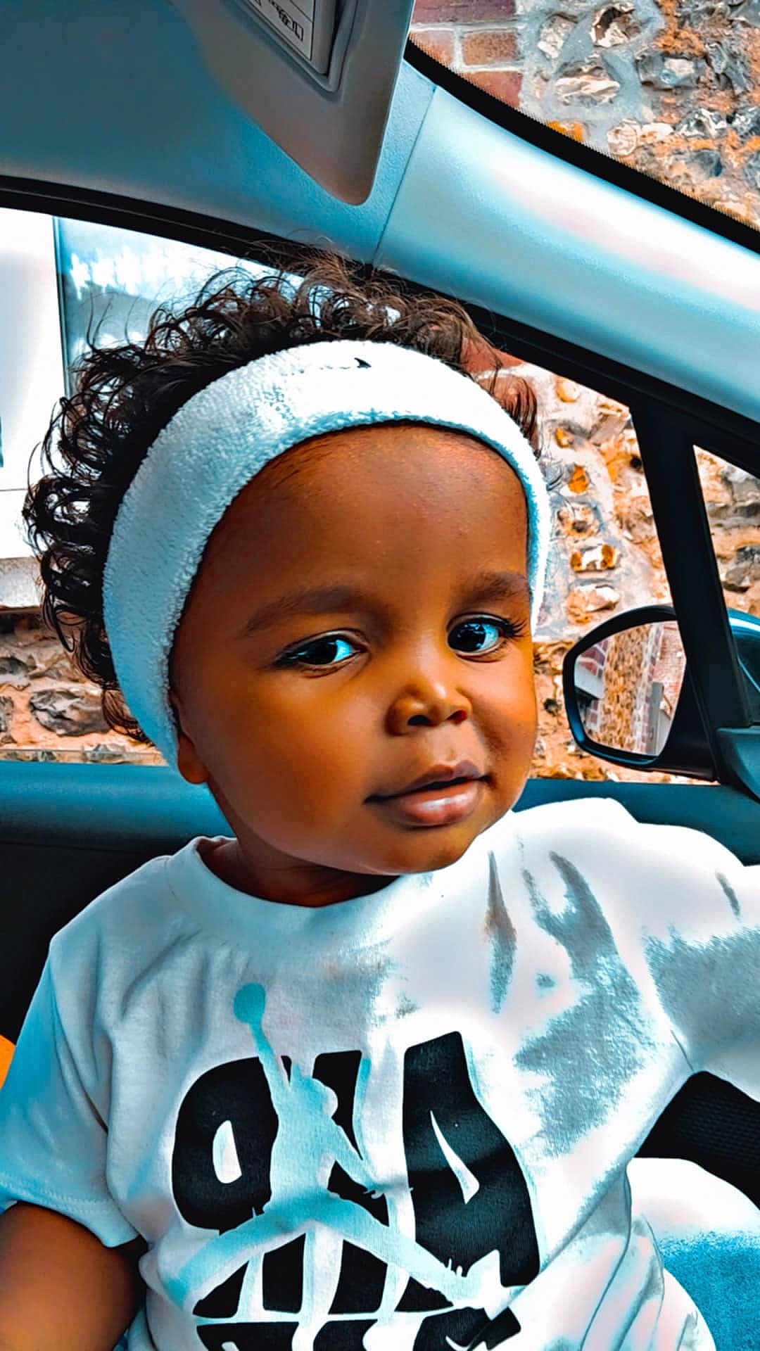 Kaïss participe au concours pour gagner de l'argent avec cette photo : toddler, child, curly_hair, headband, white_tshirt, graphic_print, car_interior, window, stone_wall, face, portrait, person, sitting, young_child, cute, indoors, casual_clothing, closeup, black_hair, expression