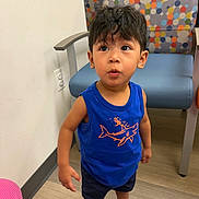 Jayden is registered to the contest to win money with this photo: child, toddler, boy, portrait, standing, wide_eyed, surprised, indoors, waiting_room, chair, colorful, polka_dot, blue_shirt, shark, shorts, sneakers, wood_floor, wall, playful, photography