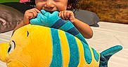 Jayden is registered to the contest to win money with this photo: child, toddler, curly_hair, smiling, face, eyes, indoors, bedroom, bed, bedding, plush_toy, stuffed_animal, fish, colorful, yellow, turquoise, portrait, sitting, playful, cozy