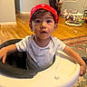 Jayden is registered to the contest to win money with this photo: baby, child, high_chair, red_cap, indoors, portrait, face, dark_hair, brown_eyes, curious, sitting, baby_clothing, white_shirt, home, hardwood_floor, rug, toys, playroom, furniture, photography