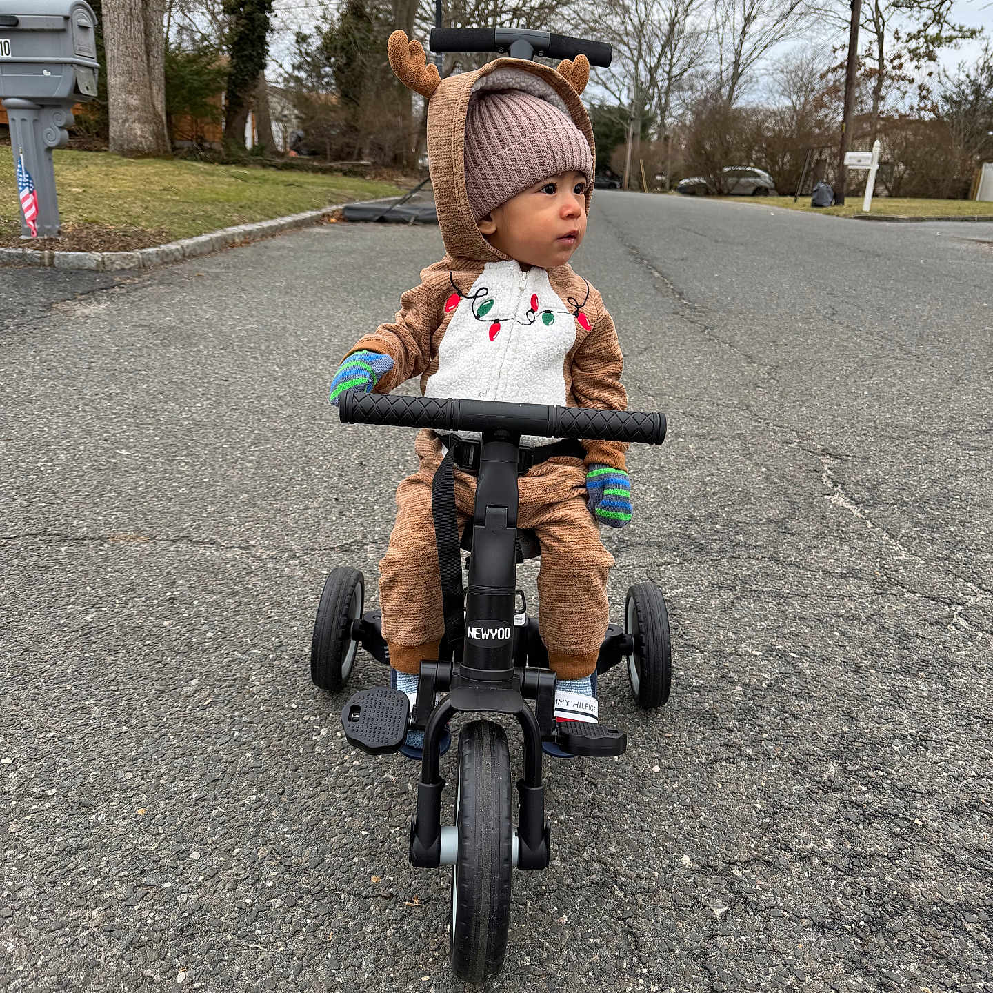 Jayden is registered to the contest to win money with this photo: toddler, child, baby, reindeer_costume, winter_clothing, beanie, outdoor, street, tricycle, ride_on_toy, vehicle, helmetless, mittens, smiling, portrait, suburban, road, trees, mailbox, daylight