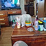 cat, white_cat, drink_cup, wooden_table, living_room, television, furniture, kitchen, bottle, spoon, mug, floor, cabinet, clutter, curious, indoor, person_leg, sitting, screen, household_items