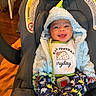 baby, smiling, car_seat, bib, hoodie, blue, yellow, dinosaur_pants, indoor, child, happy, cute, infant, clothing, seatbelt, toy, holding, face, portrait, cozy