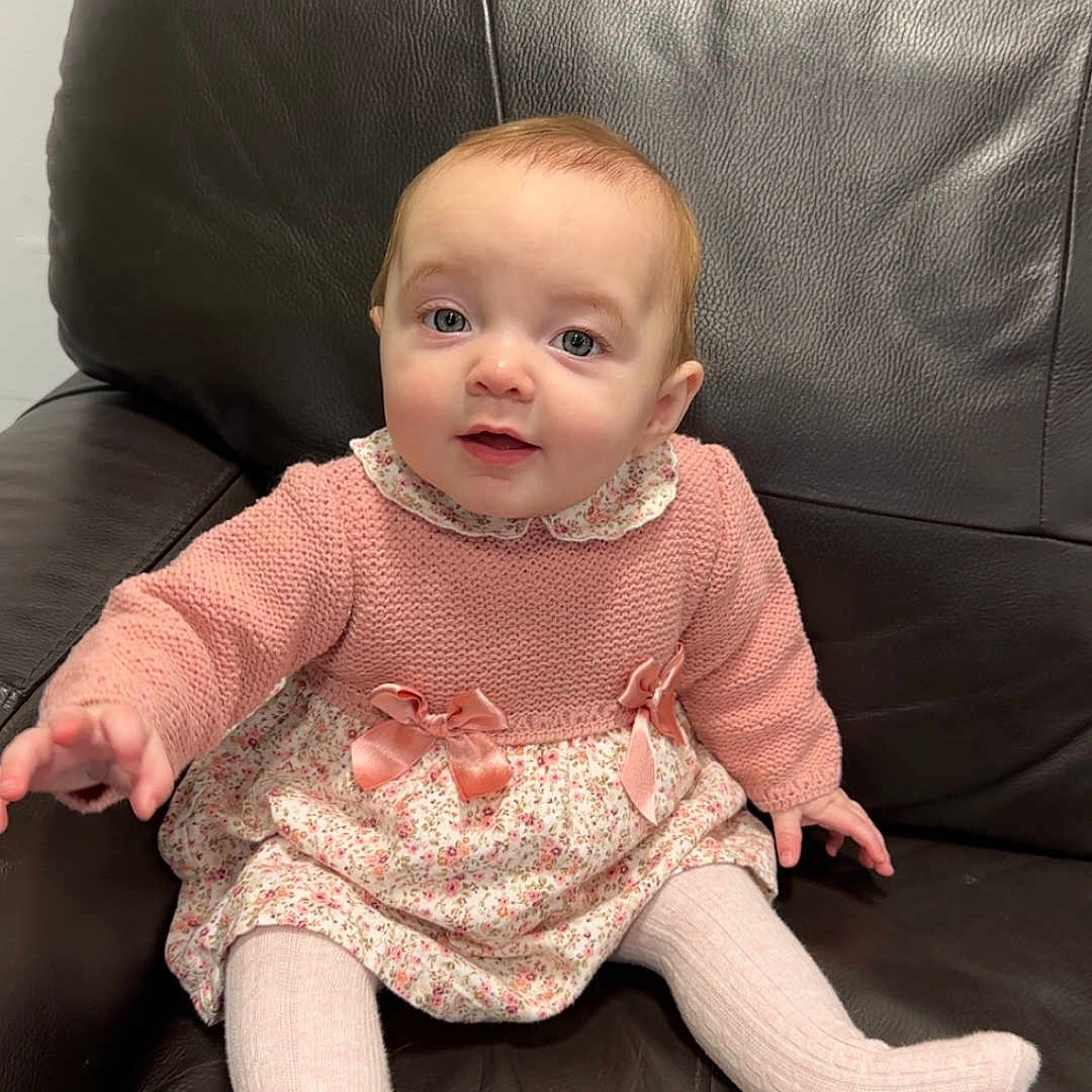 Kienna-Jo is registered to the contest to win money with this photo: baby, child, cozy, cute, expression, floral_dress, footwear, furniture, hand, indoor, infant, knitted, leather_couch, person, pink_sweater, portrait, sitting, smiling, tights, young_child