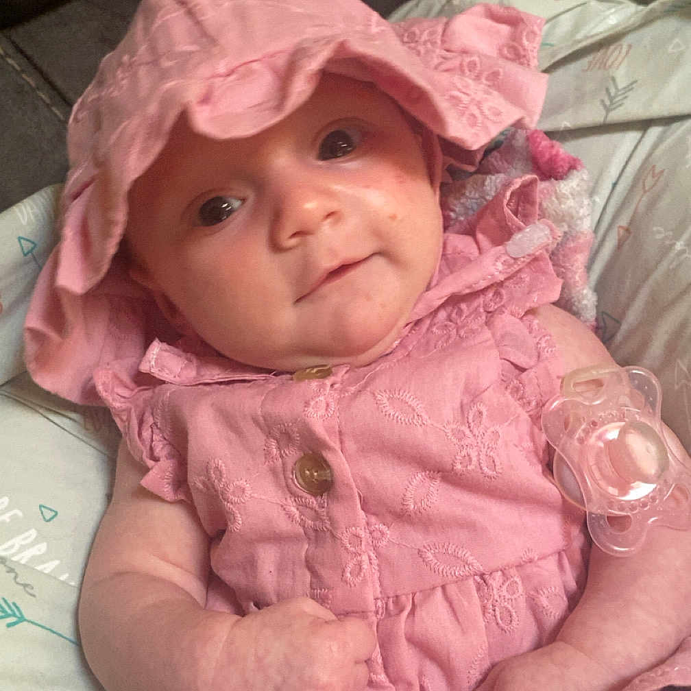 Vayda is registered to the contest to win money with this photo: adorable, baby, blanket, child, closeup, clothing, comfortable, cute, face, hands, indoors, infant, newborn, pacifier, pink_dress, portrait, resting, smiling, soft_lighting, sunhat