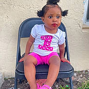 Skai is registered to the contest to win money with this photo: toddler, child, girl, sitting, chair, outdoor, pink_shorts, pink_sandals, white_shirt, number_one, hair_puffs, curly_hair, expression, looking_away, concrete_wall, grass, rocks, folding_chair, casual_clothing, daylight