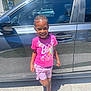 Leah joined the competition — help win amazing prizes! child, smiling, standing, braided_hair, pink_shirt, graphic_tshirt, shorts, pink_sneakers, car, black_car, outdoor, sunny, daytime, sidewalk, reflection, happy, person, clothing, vehicle, sky