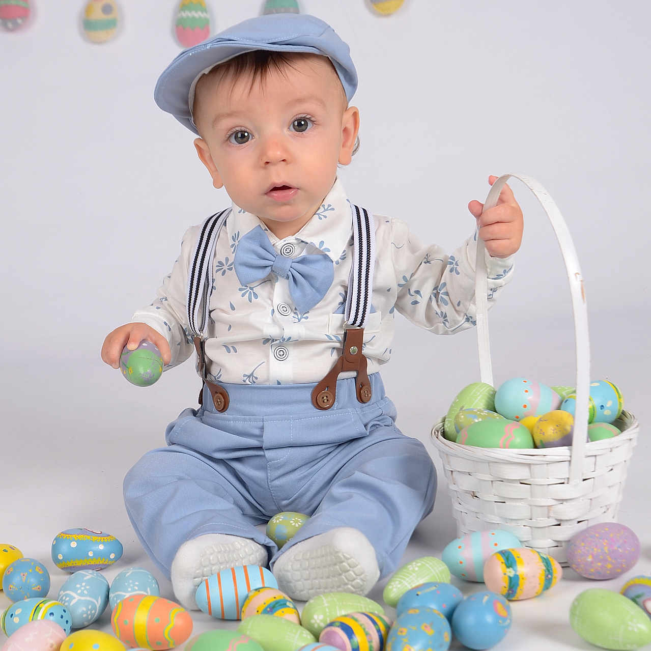 Rocco is registered to the contest to win money with this photo: baby, child, easter_eggs, basket, blue_clothing, cap, suspenders, bow_tie, studio_background, colorful, decorations, holiday, celebration, sitting, cute, portrait, indoor, festive, toys, smiling