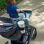 toddler, stroller, child, outdoor, path, trees, greenery, cup_holder, toy, walking, person, footwear, crocs, shade, sunlight, side_view, curly_hair, casual_clothing, nature, daytime