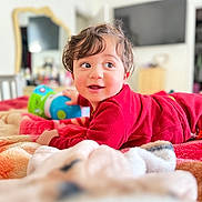 Issa is registered to the contest to win money with this photo: toddler, child, baby, red_clothing, bed, blanket, toy, smiling, face, eyes, hair, indoor, furniture, mirror, television, room, curly_hair, happy, lying_down, playful