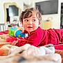 toddler, child, baby, red_clothing, bed, blanket, toy, smiling, face, eyes, hair, indoor, furniture, mirror, television, room, curly_hair, happy, lying_down, playful