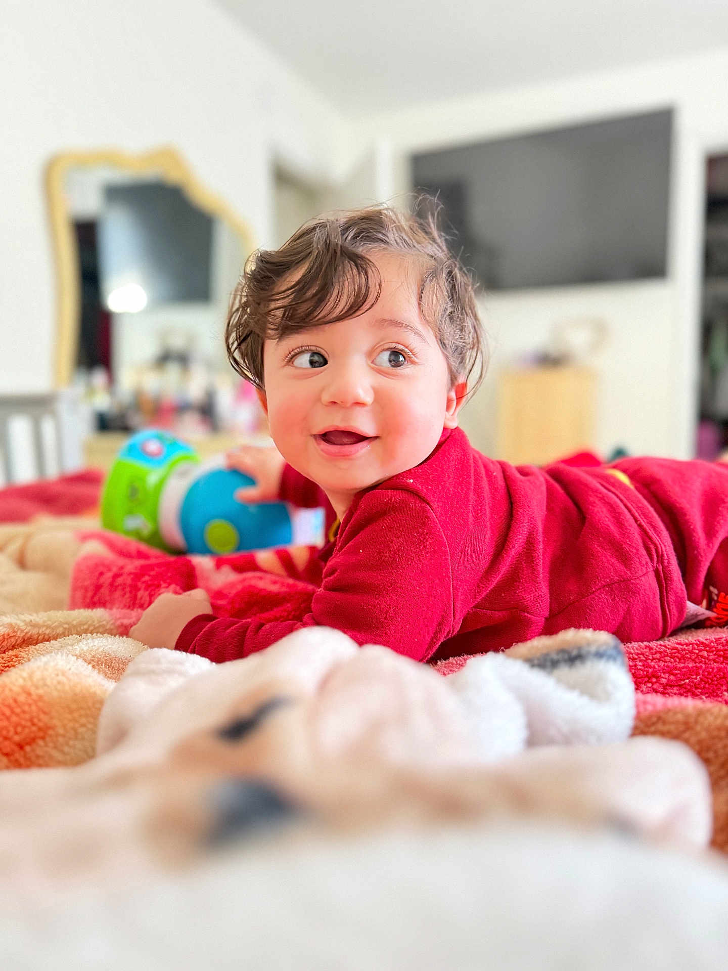 Issa is registered to the contest to win money with this photo: toddler, child, baby, red_clothing, bed, blanket, toy, smiling, face, eyes, hair, indoor, furniture, mirror, television, room, curly_hair, happy, lying_down, playful