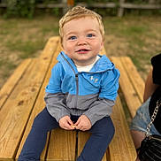 George joined the competition — help win amazing prizes! toddler, child, smiling, blue_eyes, jacket, pants, sandals, picnic_table, outdoor, grass, wood, fence, person, snack, casual_clothing, happy, sitting, portrait, daylight, nature