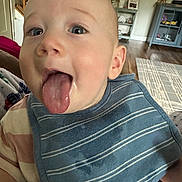 Milo joined the competition — help win amazing prizes! baby, tongue, bib, striped_clothing, indoor, living_room, furniture, carpet, wood_floor, cabinet, plant, curious, close_up, happy, child, person, face, head, smile, playful