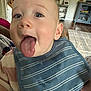 baby, tongue, bib, striped_clothing, indoor, living_room, furniture, carpet, wood_floor, cabinet, plant, curious, close_up, happy, child, person, face, head, smile, playful