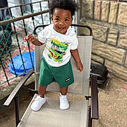 Asaan H is registered to the contest to win money with this photo: toddler, child, smiling, standing, chair, outdoor, white_tshirt, green_shorts, white_sneakers, curly_hair, fence, brick_wall, concrete, metal_armrest, happy, person, portrait, daylight, casual_clothing, young_child