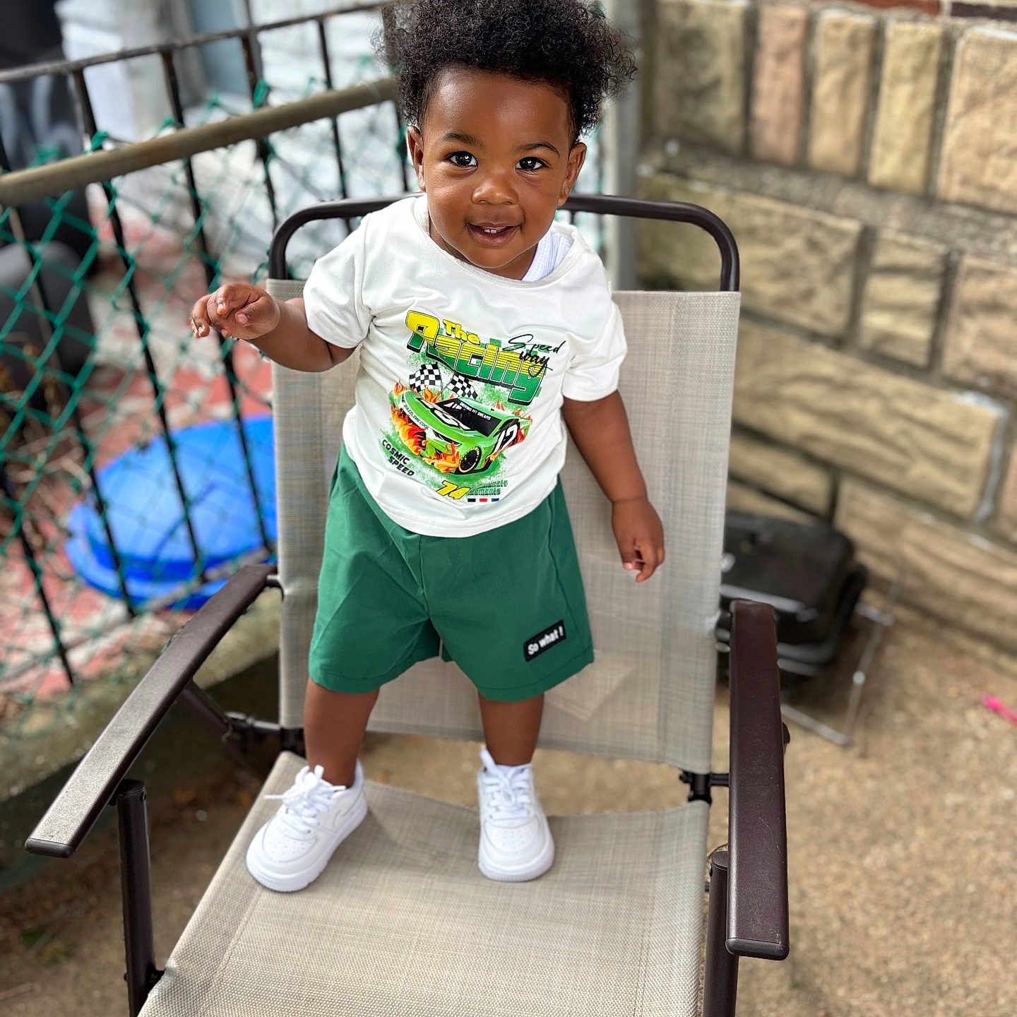 Asaan H is registered to the contest to win money with this photo: toddler, child, smiling, standing, chair, outdoor, white_tshirt, green_shorts, white_sneakers, curly_hair, fence, brick_wall, concrete, metal_armrest, happy, person, portrait, daylight, casual_clothing, young_child