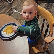 Jason is registered to the contest to win money with this photo: toddler, child, macaroni_and_cheese, fork, wooden_chair, dining_table, plate, kitchen, flooring, green_sweater, blue_pants, indoor, food, meal, person, curious_expression, sitting, wooden_furniture, young_child, family