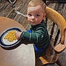 toddler, child, macaroni_and_cheese, fork, wooden_chair, dining_table, plate, kitchen, flooring, green_sweater, blue_pants, indoor, food, meal, person, curious_expression, sitting, wooden_furniture, young_child, family