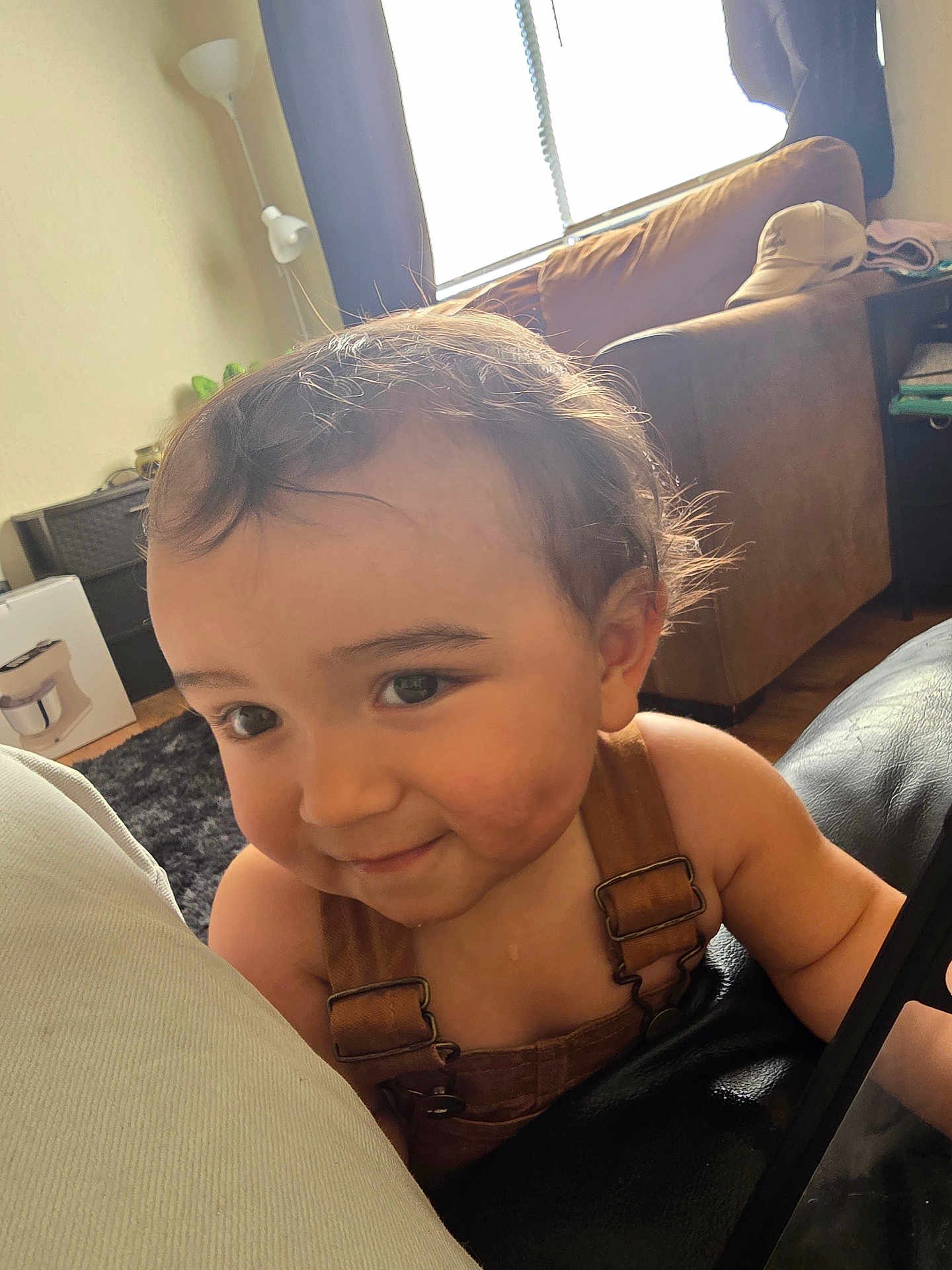Makai is registered to the contest to win money with this photo: toddler, child, smiling, face, overalls, indoors, living_room, window, curtains, sofa, lamp, floor, carpet, person, hair, skin, cheeks, happy, casual, closeup