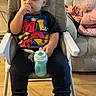 child, toddler, high_chair, sippy_cup, boots, clothing, indoors, floor, couch, furniture, person, face, hand, sitting, wooden_floor, casual, home, young_child, cute, thinking
