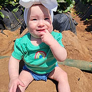 Harper is registered to the contest to win money with this photo: baby, child, headband, bow, green_shirt, rainbow, smile, fingers_in_mouth, shorts, sand, outdoor, sunny, plants, strawberries, field, nature, portrait, happy, sitting, person
