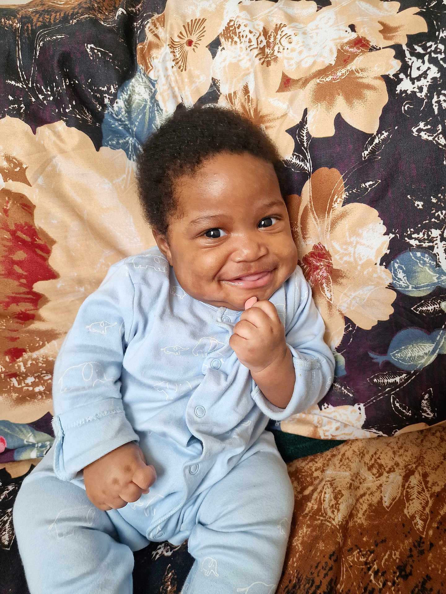 Aku joined the competition — help win amazing prizes! baby, smiling, blue_onesie, elephant_print, sitting, hand_fist, curly_hair, floral_blanket, happy, cheeky, cute, infant, portrait, indoor, child, furniture, cozy, playful, face, young