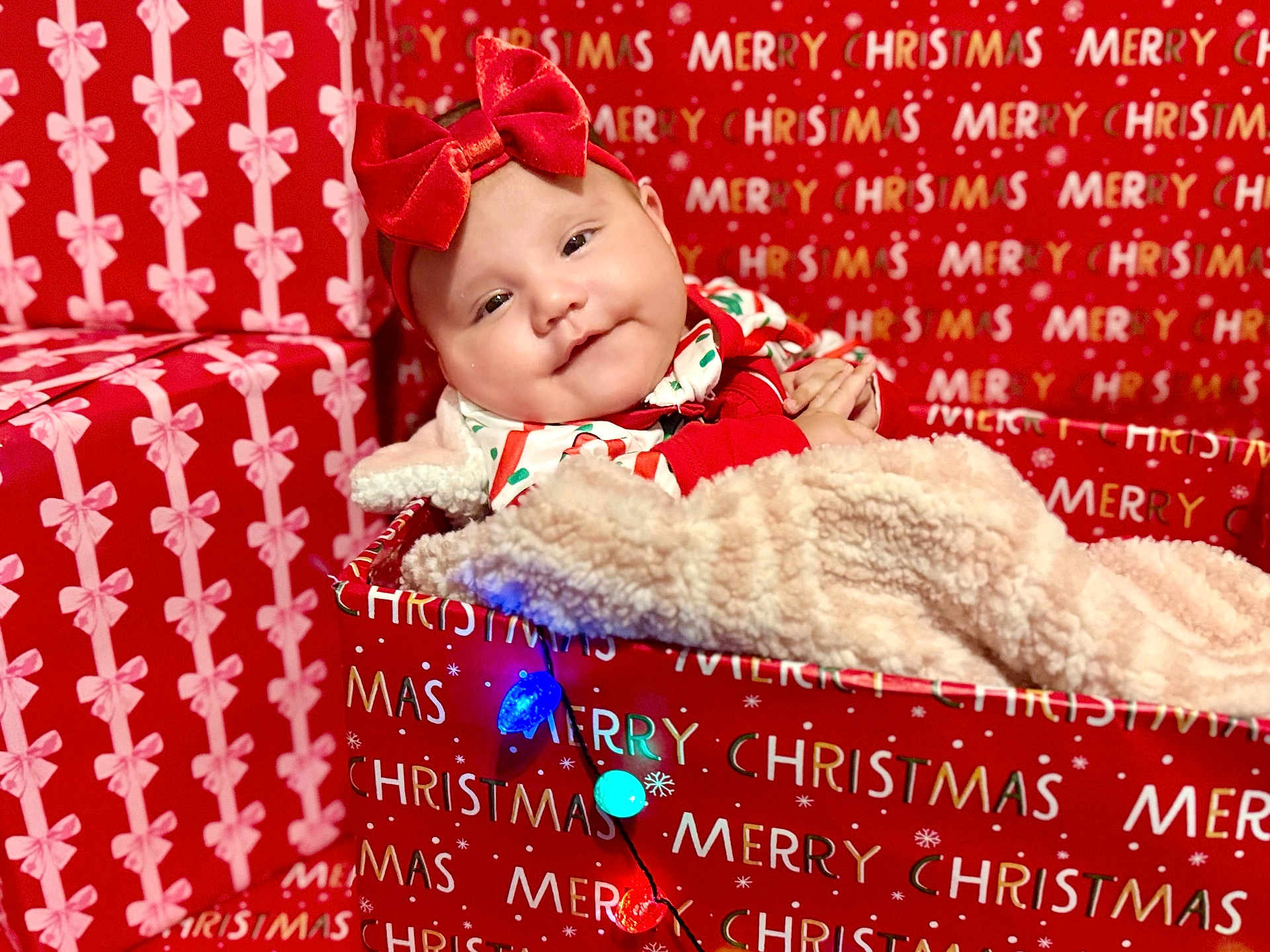 Iris is registered to the contest to win money with this photo: baby, red_bow, headband, christmas, holiday_lights, gift_box, blanket, wrapping_paper, festive, smiling, cute, infant, cozy, celebration, indoors, red_clothing, soft_texture, portrait, happy, seasonal
