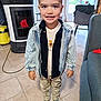 child, boy, indoor, smiling, standing, denim_jacket, striped_socks, pants, shirt, floor_tiles, living_room, window, chair, stove, natural_light, casual_clothing, young_child, happy, person, home