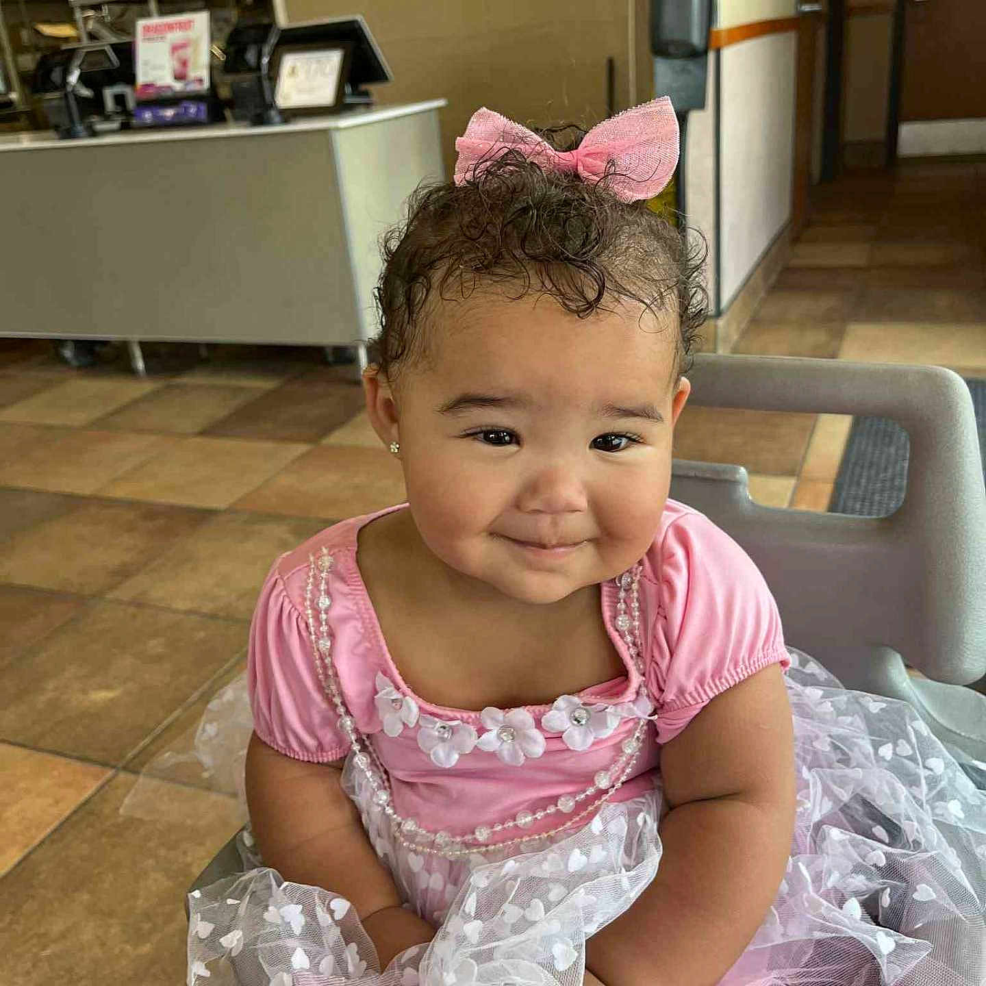 Honesty is registered to the contest to win money with this photo: baby, bow, child, curly_hair, cute, dress, earrings, fashion, floor_tiles, happy, high_chair, indoor, person, pink_dress, portrait, restaurant, seat, smile, sweet, toddler
