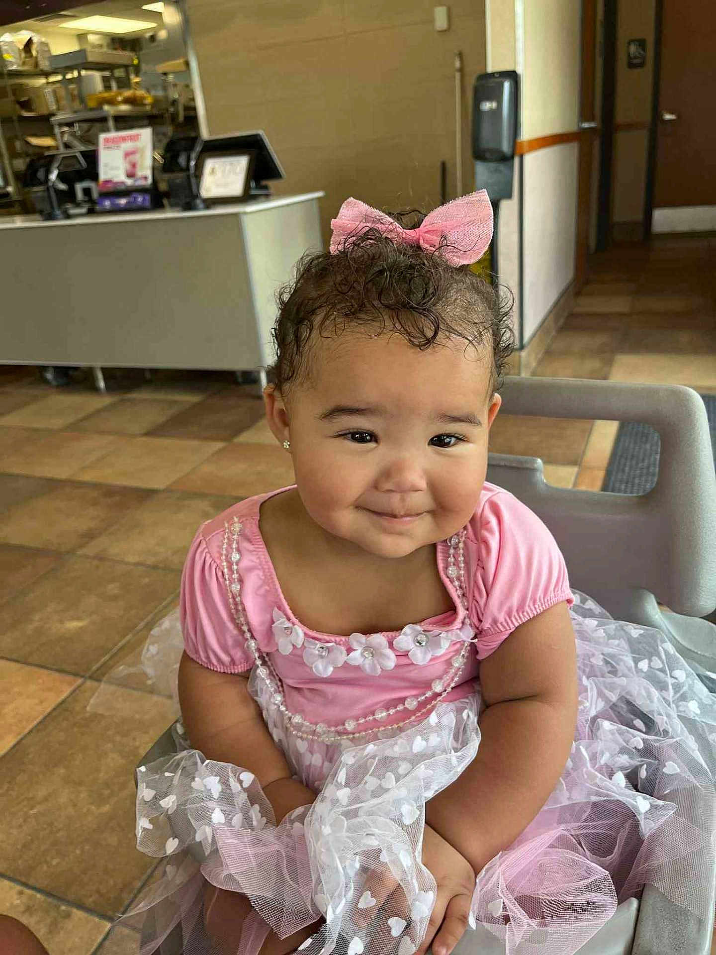 Honesty is registered to the contest to win money with this photo: toddler, child, pink_dress, bow, smile, curly_hair, high_chair, restaurant, floor_tiles, earrings, cute, happy, portrait, indoor, person, fashion, baby, seat, dress, sweet