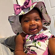 Navonni is registered to the contest to win money with this photo: baby, child, smiling, hat, floral, clothing, indoor, person, cute, portrait, happy, infant, skin, hand, face, headwear, purple, fabric, holding, closeup