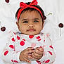 baby, child, red_bow, headband, cherry_pattern, long_sleeve_shirt, white_blanket, fruit, cherries, hand, cute, infant, lying_down, playful, curious_expression, skin, face, portrait, one_person, indoors