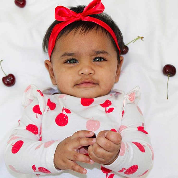 Amyra is registered to the contest to win money with this photo: baby, cherries, cherry_pattern, child, curious_expression, cute, face, fruit, hand, headband, indoors, infant, long_sleeve_shirt, lying_down, one_person, playful, portrait, red_bow, skin, white_blanket