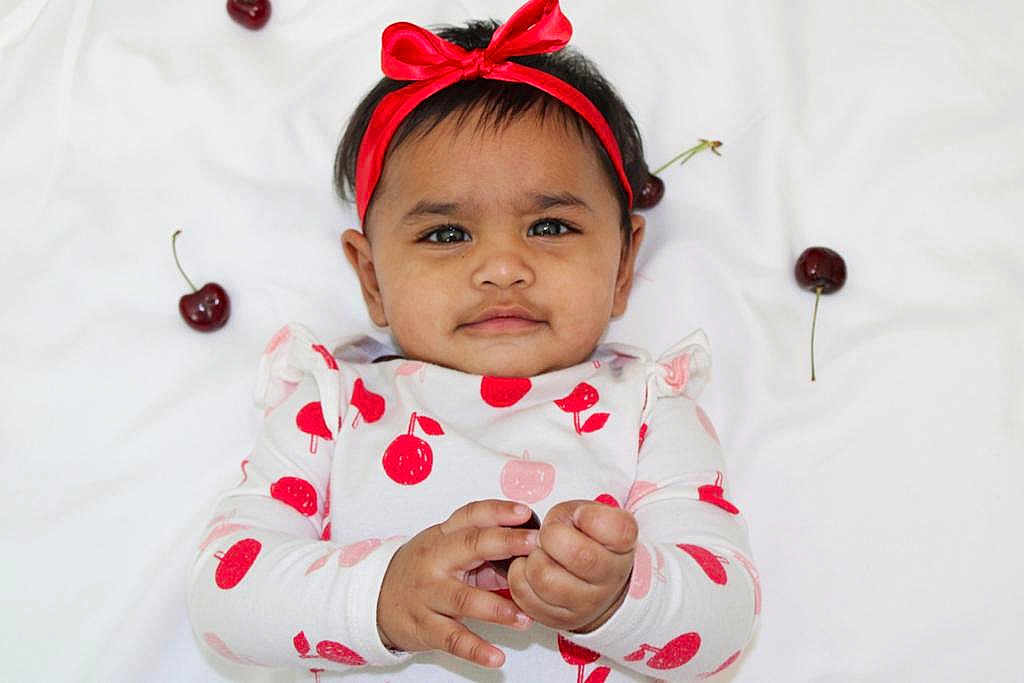 Amyra is registered to the contest to win money with this photo: baby, child, red_bow, headband, cherry_pattern, long_sleeve_shirt, white_blanket, fruit, cherries, hand, cute, infant, lying_down, playful, curious_expression, skin, face, portrait, one_person, indoors