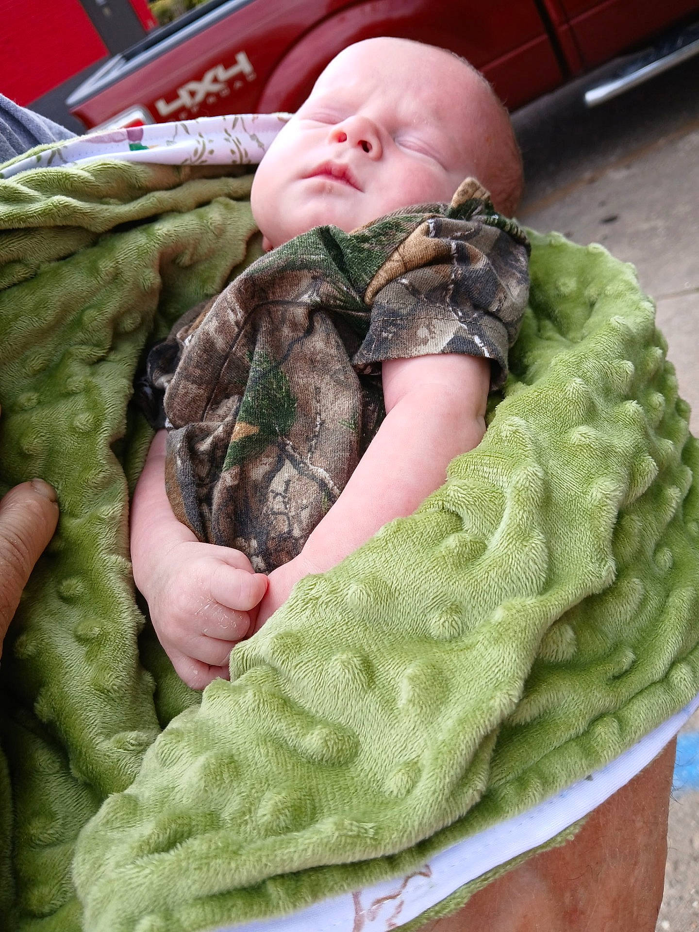 William is registered to the contest to win money with this photo: baby, blanket, camo_clothing, child, closeup, fabric, face, green_blanket, hand, infant, outdoor, peaceful, person, portrait, red_truck, sleep, sleeping, texture, vehicle, wrapped