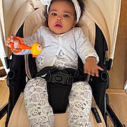 Ghalia participe au concours pour gagner de l'argent avec cette photo : baby, stroller, headband, toy, onesie, fish_pattern, cardigan, indoor, floor, wooden_floor, pet_bowl, curly_hair, infant, child, seatbelt, baby_clothing, black_hair, cute, looking_at_camera, baby_toy