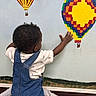 toddler, child, denim_overalls, white_shirt, curly_hair, wall_mural, hot_air_balloon, colorful, indoor, art, painting, exploration, imagination, back_view, hand, baby, crawling, playful, cute, young_child