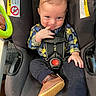 baby, child, car_seat, plaid_shirt, blue_eyes, brown_hair, finger_in_mouth, booties, smile, cute, infant, seatbelt, clothing, indoor, child_safety, toddler, playful, person, portrait, seat