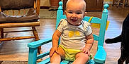 Josie is registered to the contest to win money with this photo: baby, rocking_chair, smile, indoor, wooden_chair, floor_rug, potted_plant, door, clothing, baby_clothes, dog_tail, wood_floor, furniture, child, happy, person, living_room, table, window, backpack