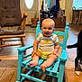 baby, rocking_chair, smile, indoor, wooden_chair, floor_rug, potted_plant, door, clothing, baby_clothes, dog_tail, wood_floor, furniture, child, happy, person, living_room, table, window, backpack
