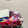 Josie is registered to the contest to win money with this photo: child, toddler, wagon, hat, purple_clothing, towel, outdoor, parking_lot, car, smiling, sunny, asphalt, wheel, red_wagon, person, baby, play, happy, summer, casual