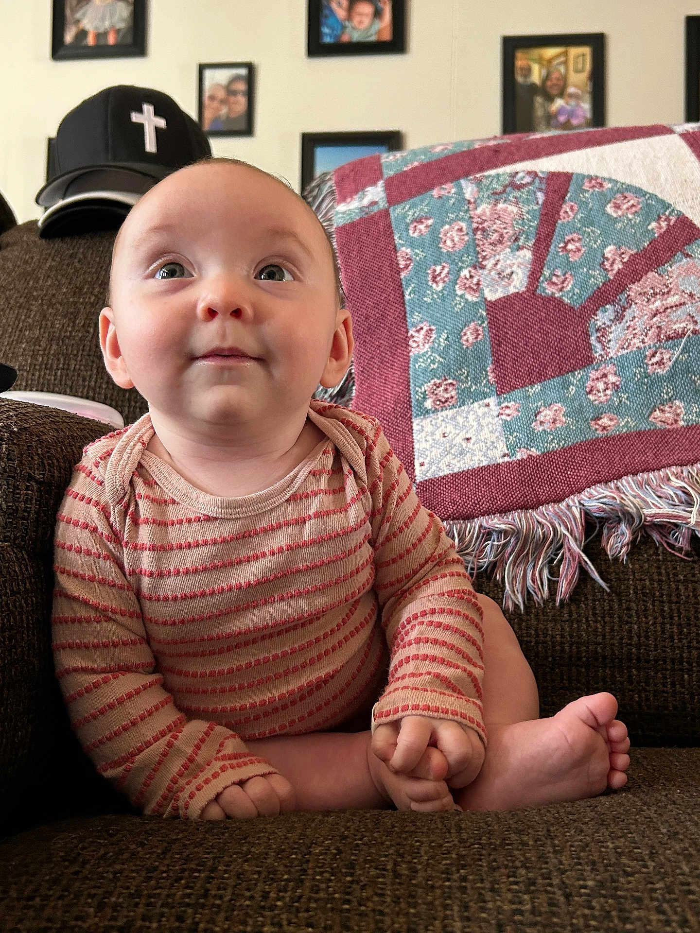 Valerie is registered to the contest to win money with this photo: baby, child, striped_onesie, couch, quilt, patchwork, family_photos, wall, indoor, seated, curious, hands, feet, head, face, expression, furniture, home, cozy, portrait
