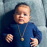 Yaya participe au concours pour gagner de l'argent avec cette photo : baby, infant, child, face, portrait, clothing, necklace, gold_jewelry, blue_shirt, hand, fingers, lying_down, texture, gray_background, cute, expression, young, skin, person, indoors